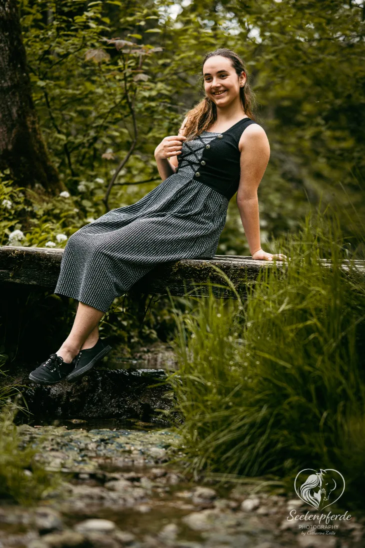 Frau in Tracht sitzt auf einem Holzsteg über einem Bach im grünen Wald